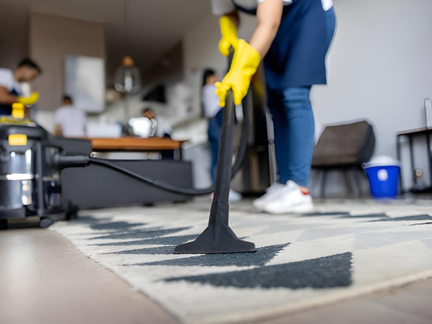Person wearing yellow gloves and a blue apron vacuuming a rug